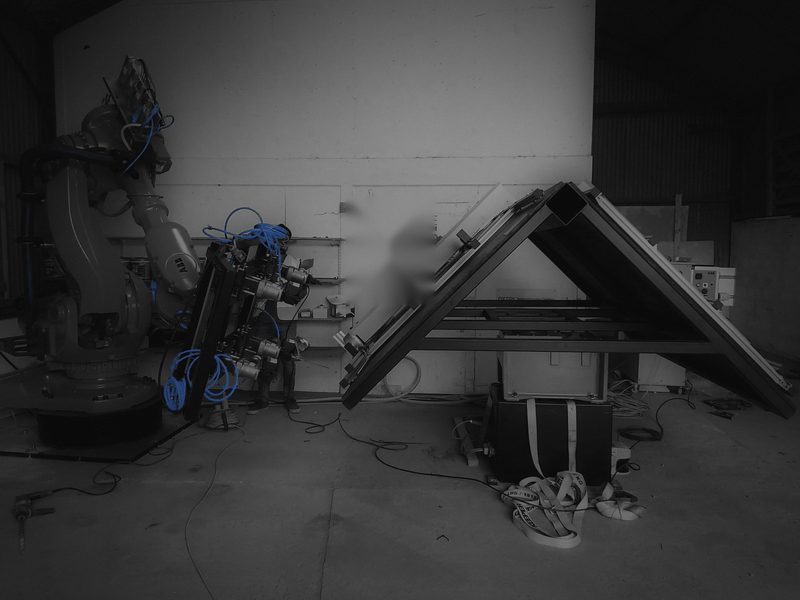 Industrial robot with a multi-tool end effector beside a tilting turntable rig, dark-graded